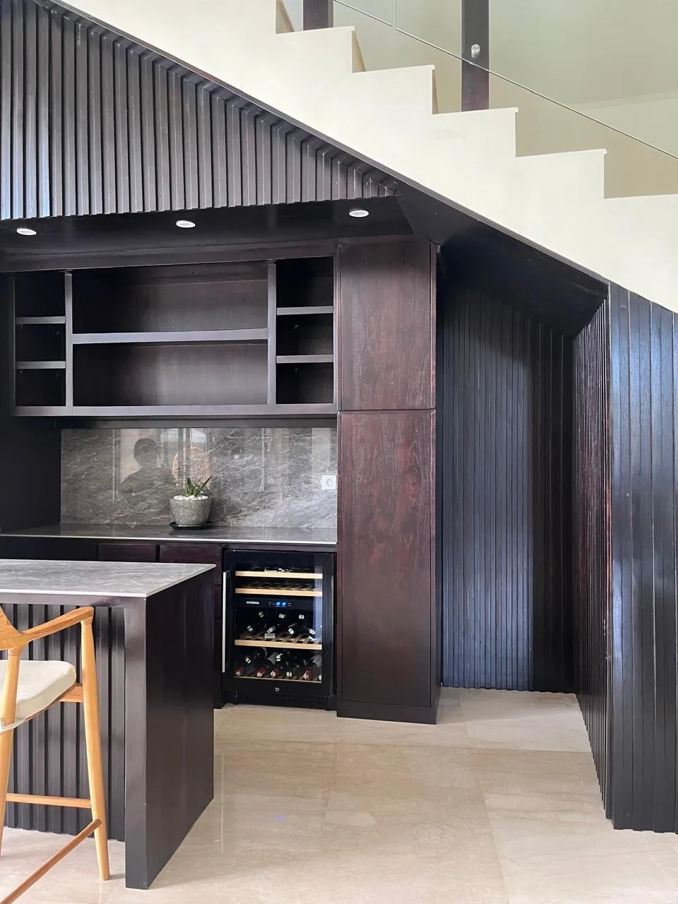 A fluted dark-timber bar unit with open shelving and a stone counter, built under a staircase