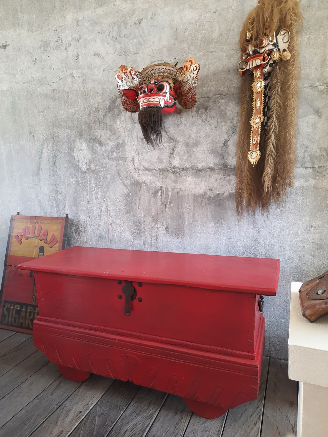 A red-painted antique Balinese chest below a carved Barong mask on a raw concrete wall