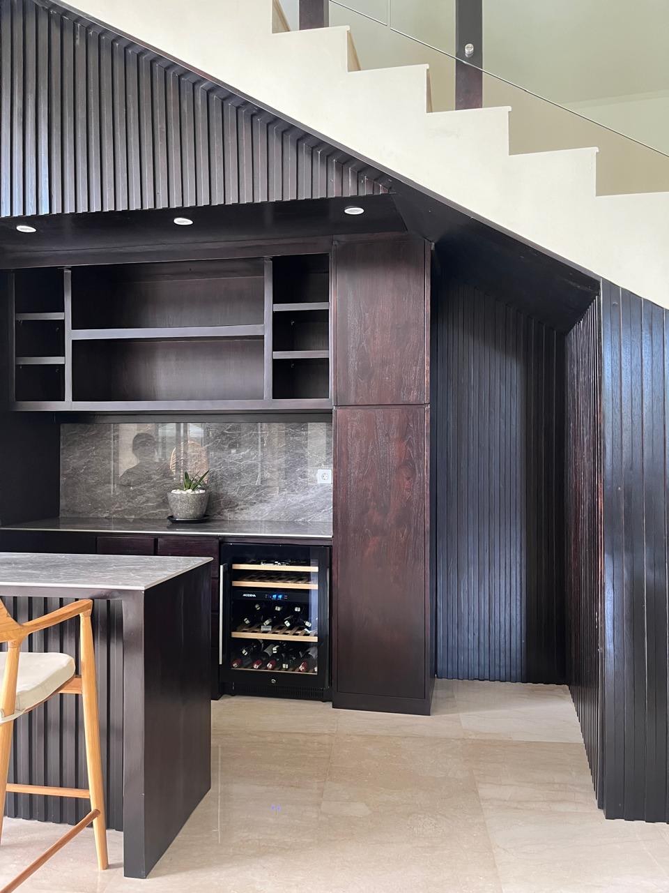 A fluted dark-timber bar unit with open shelving and a stone counter, built under a staircase
