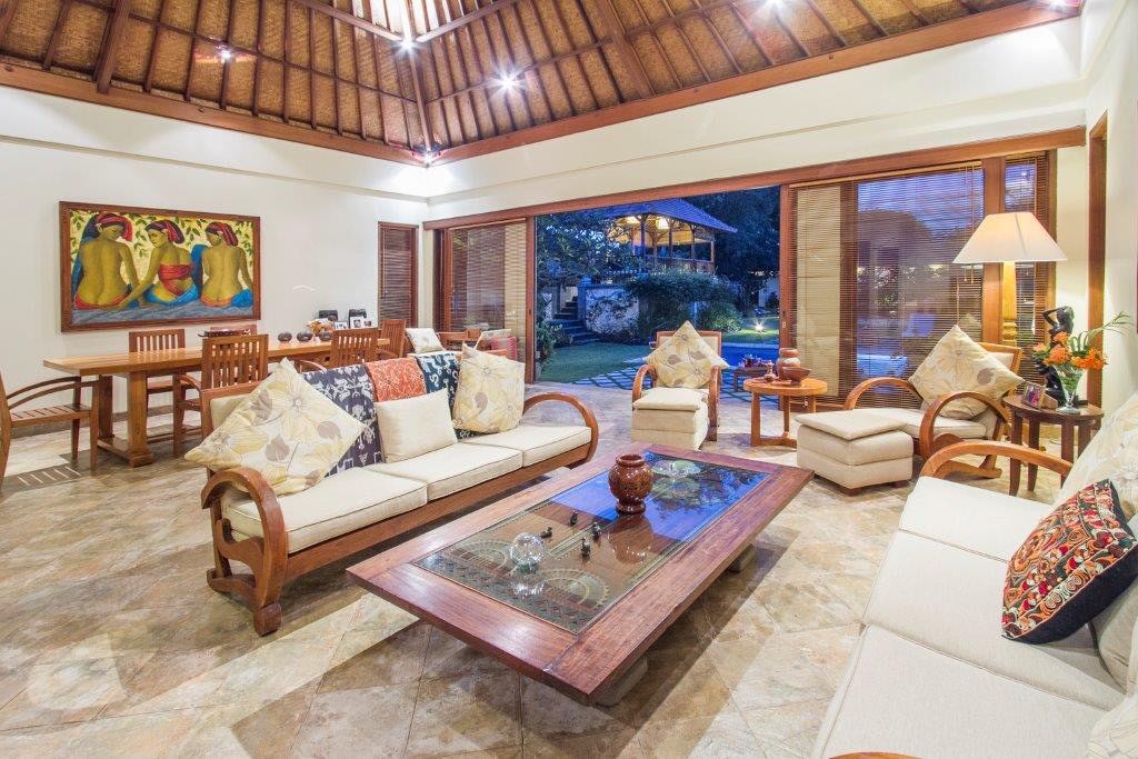 An open-sided living pavilion under an alang-alang roof, with teak sofas, a glass-topped carved coffee table and a long dining table