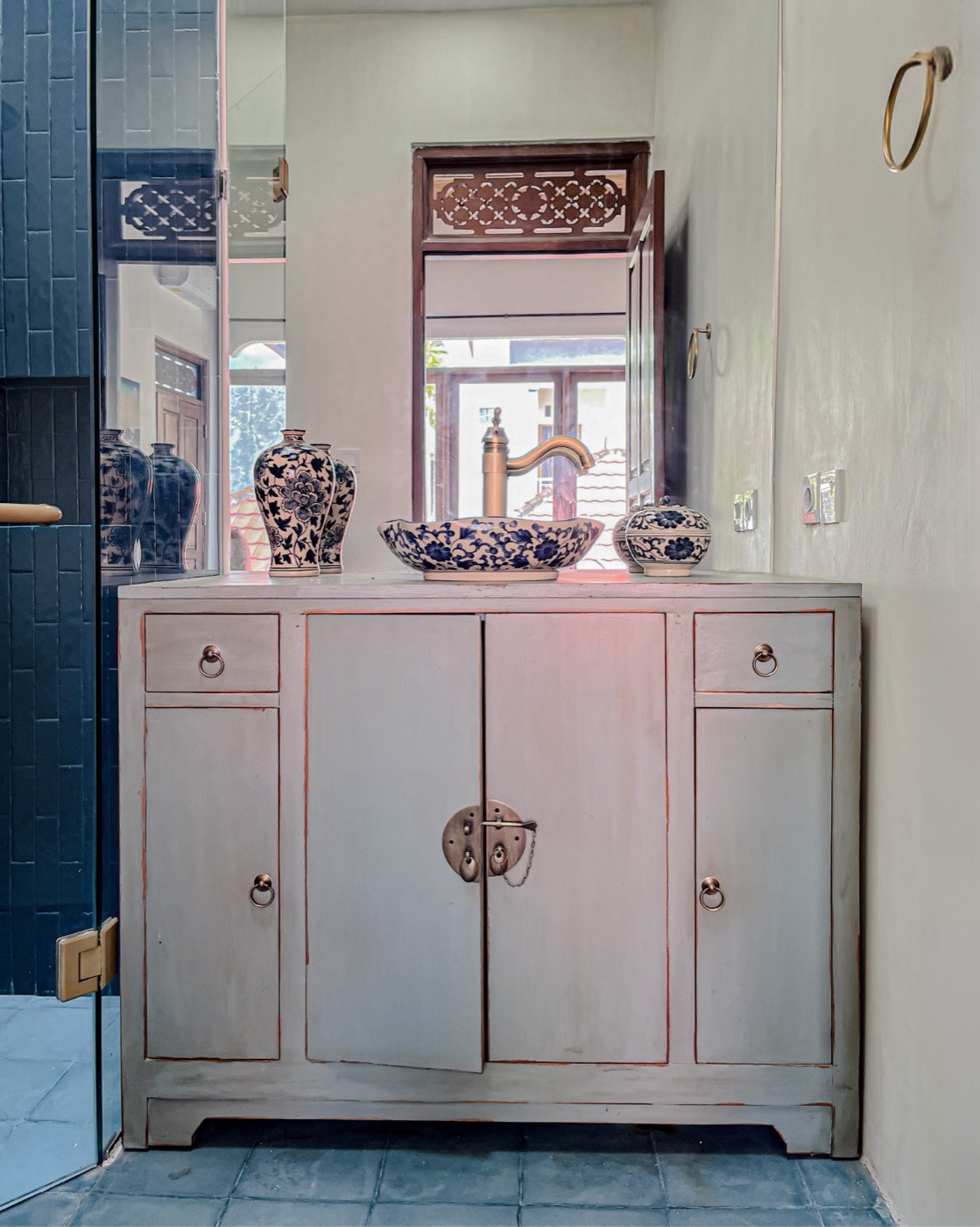 A pale grey Chinese-style cabinet with brass ring pulls, used as a bathroom vanity with blue-and-white ceramics