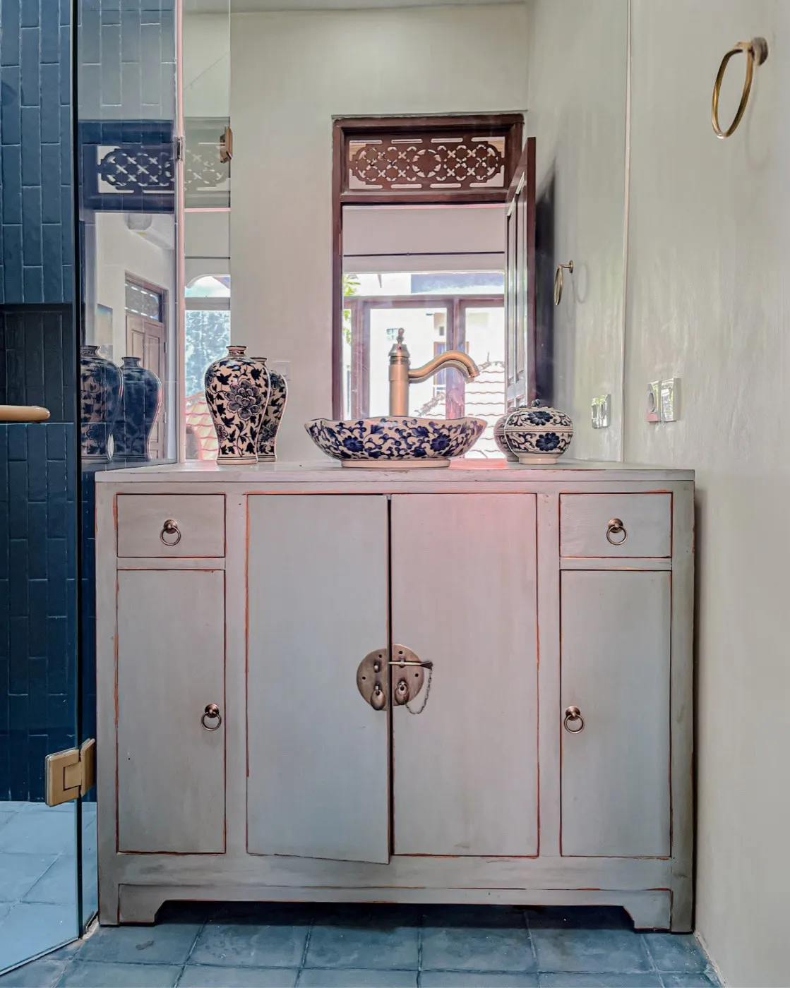 A pale grey Chinese-style cabinet with brass ring pulls, used as a bathroom vanity with blue-and-white ceramics
